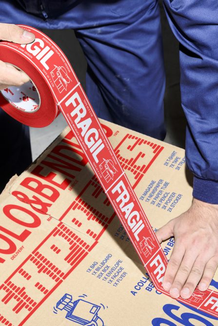 Packaging mockup featuring a person applying red Fragile tape to a cardboard box. Ideal for designers, this mockup highlights shipping and logistics design.