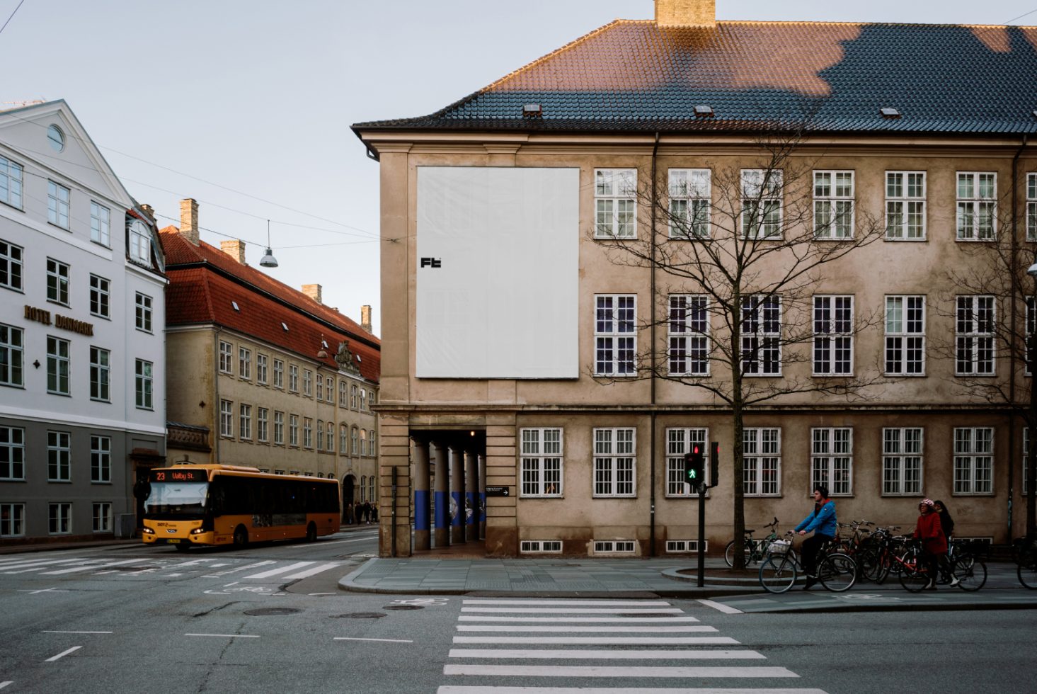 Urban street scene featuring a large billboard mockup on a historic building, with a bus passing and cyclists at the intersection on a clear day.