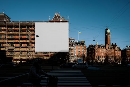 Blank large billboard on a building under renovation in a city square with a cyclist passing by in the foreground. Ideal for urban mockup and ad designs.