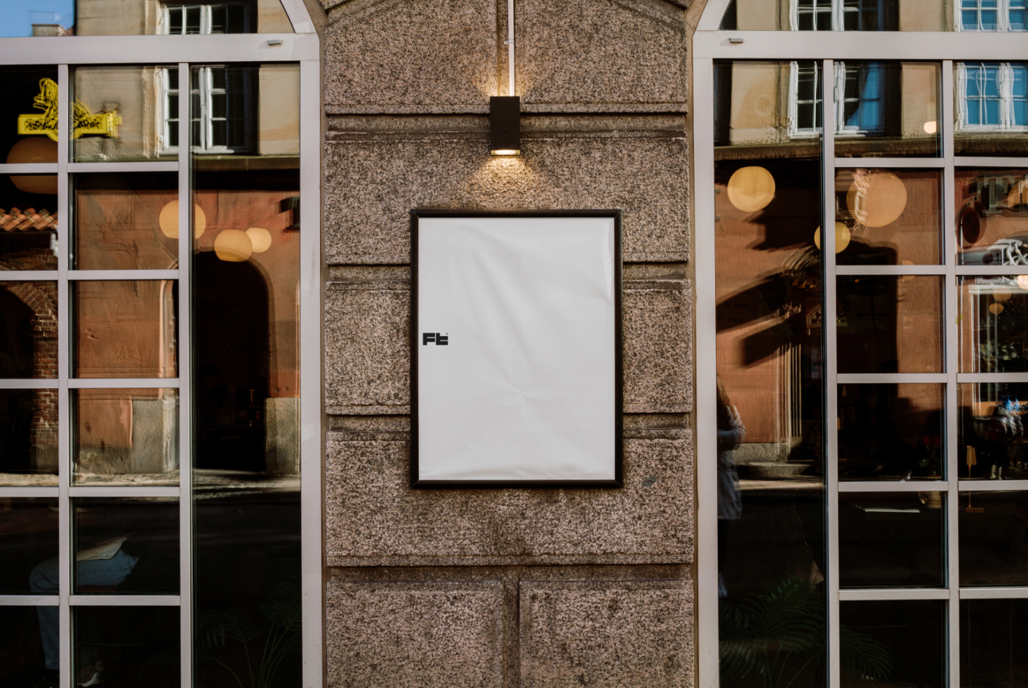 Mockup design featuring a blank poster in a black frame mounted on an outdoor stone wall with reflections from adjacent buildings and window frames.