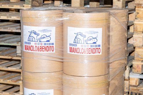 Large rolls of sticker paper labeled Manolo & Bendito 300x220 mm on stacked pallets in a warehouse. Keywords: labels, packaging, warehouse, stickers.