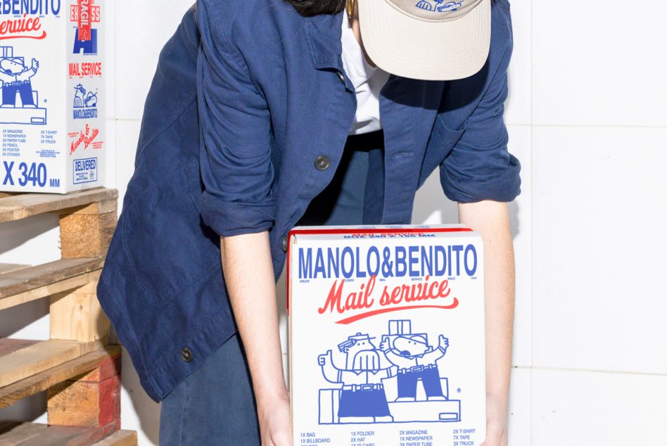 Person lifting branded Manolo & Bendito shipping box, wearing cap, blue jacket. Design features retro cartoon characters. Perfect for packaging mockup designs