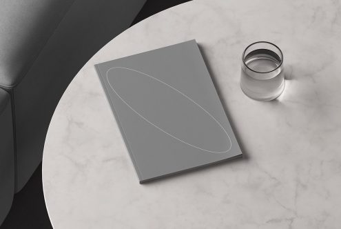 Gray book cover mockup on a marble table with a glass of water, couch in the corner ideal for showcasing book cover designs templates graphic assets for designers