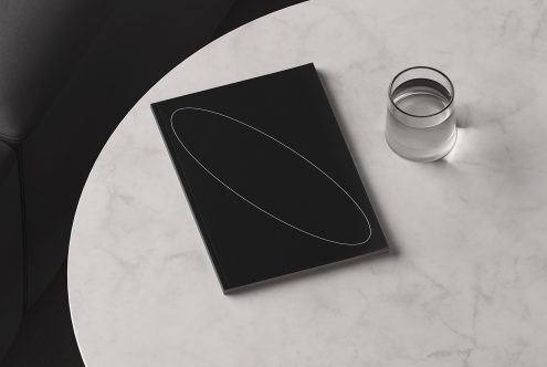 Top view mockup of a black notebook with a minimalistic design on a marble table next to a glass of water. Perfect for showcasing branding or stationery designs