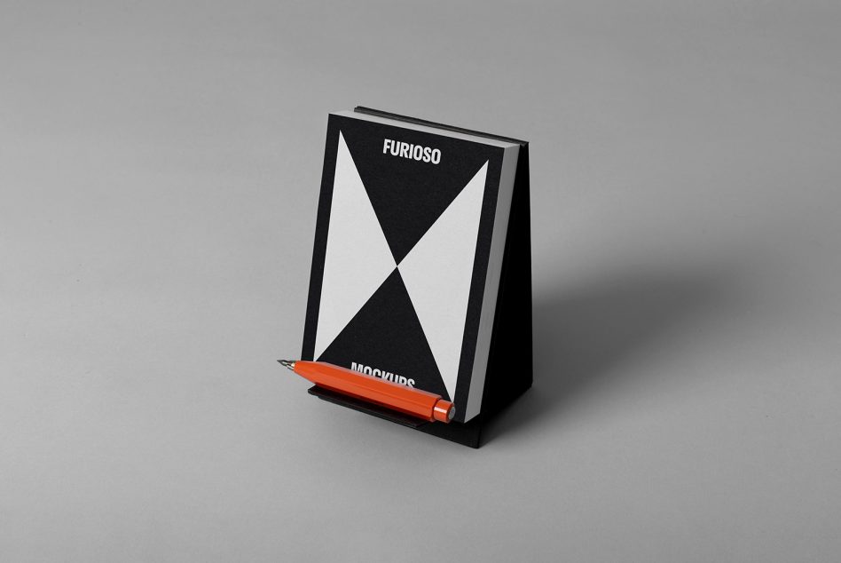 Book mockup with geometric black and white cover displayed on a stand grey backdrop orange pen beside. Perfect for showcasing print designs.