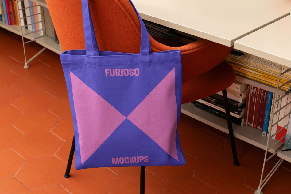 Tote bag mockup with vibrant purple and pink geometric design displayed on a chair in a modern interior setting against a bookshelf.