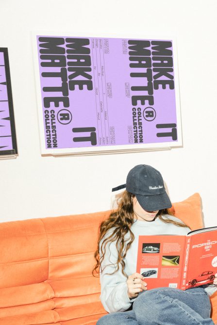 A designer wearing a cap reads a book on an orange couch; behind them is a bold typographic poster mockup in purple and black. Tags Mockups, Graphics.