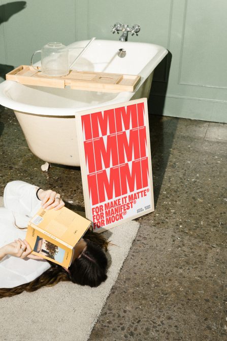 Poster mockup leaning against a bathtub with a wooden tray, next to a person laying on carpet reading a book. Keywords: Poster, Mockup, Design, Template, Graphics.