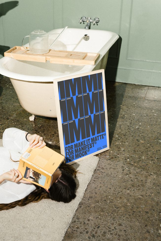 Poster mockup leaning against a bathtub with a woman reading on the floor nearby. Keywords: mockups, design assets, designer resources, templates, graphics