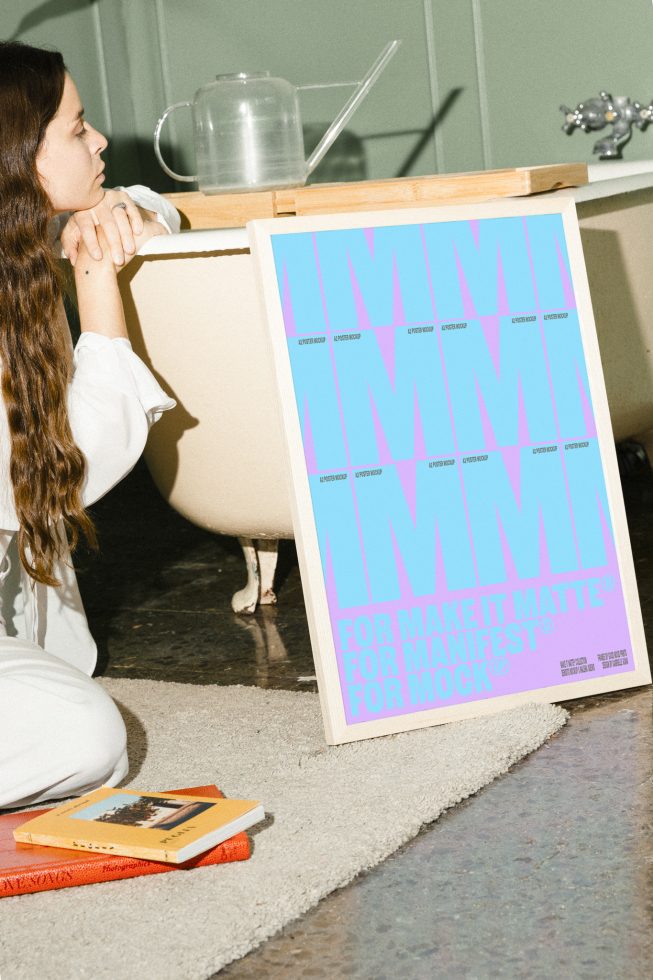 Designer sitting near bathtub with an A2 poster mockup featuring bold blue and purple text. Books on rug. Targeted for graphic designers searching mockups.