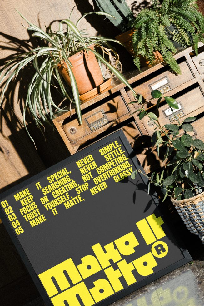 Typography design mockup featuring bold yellow text Make It Matte Never Glossy on black poster board with plants and wooden drawers in the background.