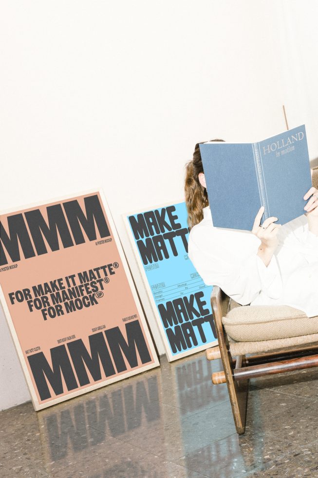 Two large posters leaning against a wall, one in beige and the other in blue, with bold black text. Person reading on a chair. Digital mockup for designers.