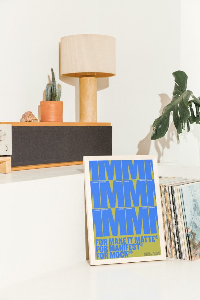 Framed typography poster mockup against wall beside shelves with cactus plant, lamp, and vinyl records. Ideal for graphic design, mockups, and templates.