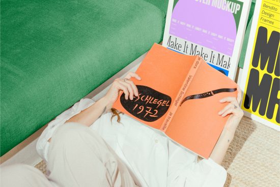 Person laying down holding an orange book with colorful posters in the background. Ideal for designers, mockups, poster design, creative assets, graphics.