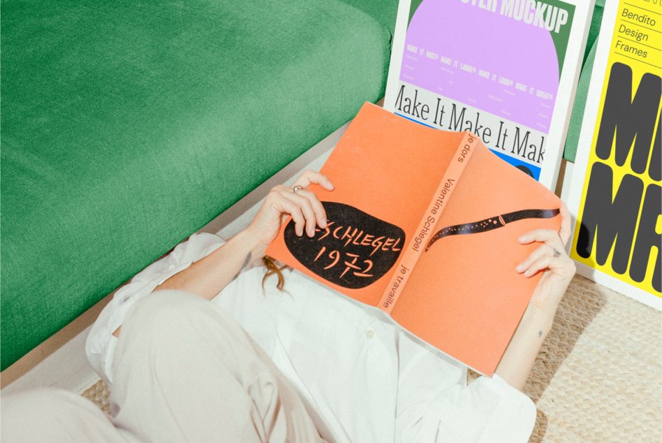 Person laying down holding an orange book with colorful posters in the background. Ideal for designers, mockups, poster design, creative assets, graphics.