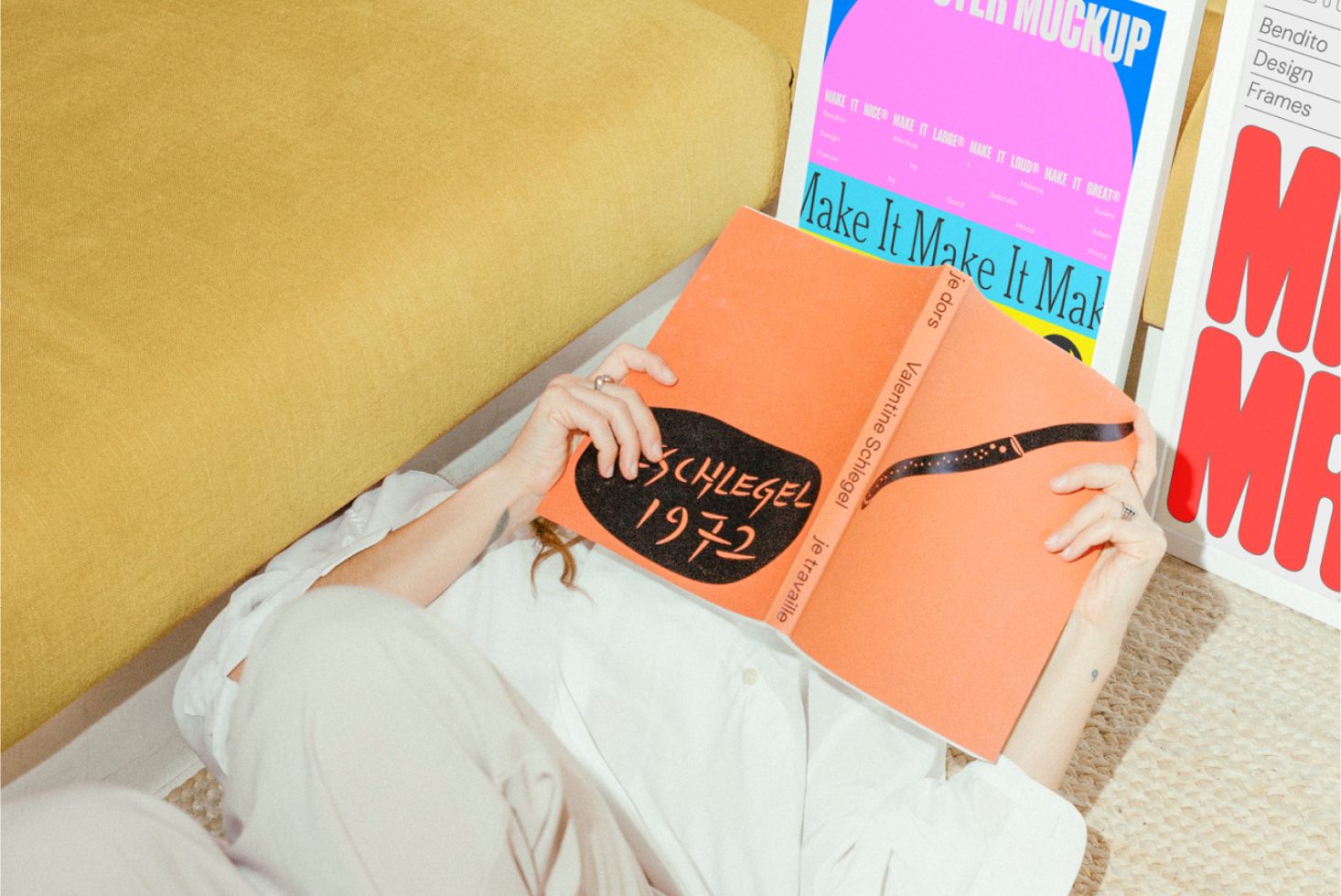 Person reading a book on retro art design while lying on a carpet next to colorful poster mockups and fonts for packaging and graphic design projects.