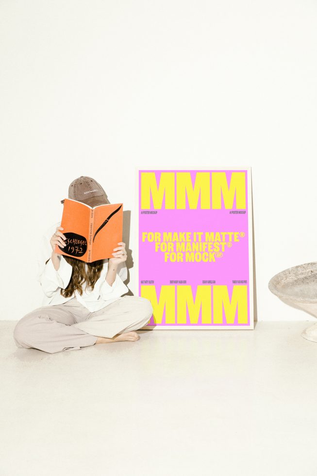 A person sitting on the floor reading an orange book with a large pink and yellow poster mockup beside them ideal for designers and creatives seeking mockup assets