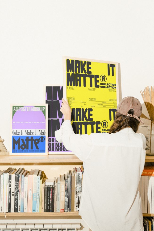 Designer placing colorful matte A3 poster mockups on a shelf filled with books ideal for showcasing design templates mockups in a creative studio setup.