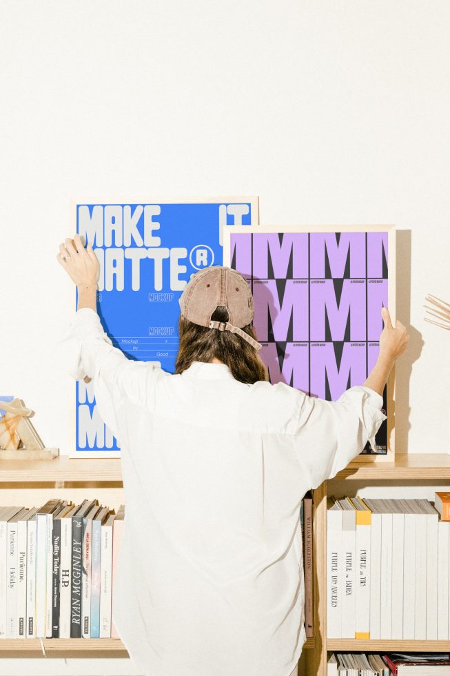 Designer hanging framed mockups in creative workspace. Shelves with books in background. Use this to showcase graphic design or typography projects.