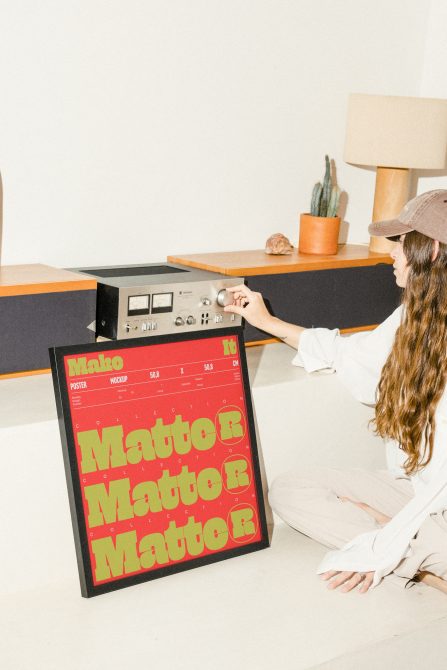 Woman adjusting vintage stereo beside framed poster mockup with bold typography in a minimalistic room with cactus and lamp in background.