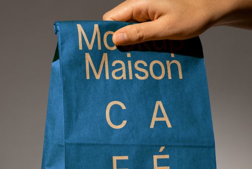 Hand holding a blue paper bag with the text Mockup Maison Café. Ideal for branding or packaging mockups. Keywords: packaging mockup, graphic design, digital asset.