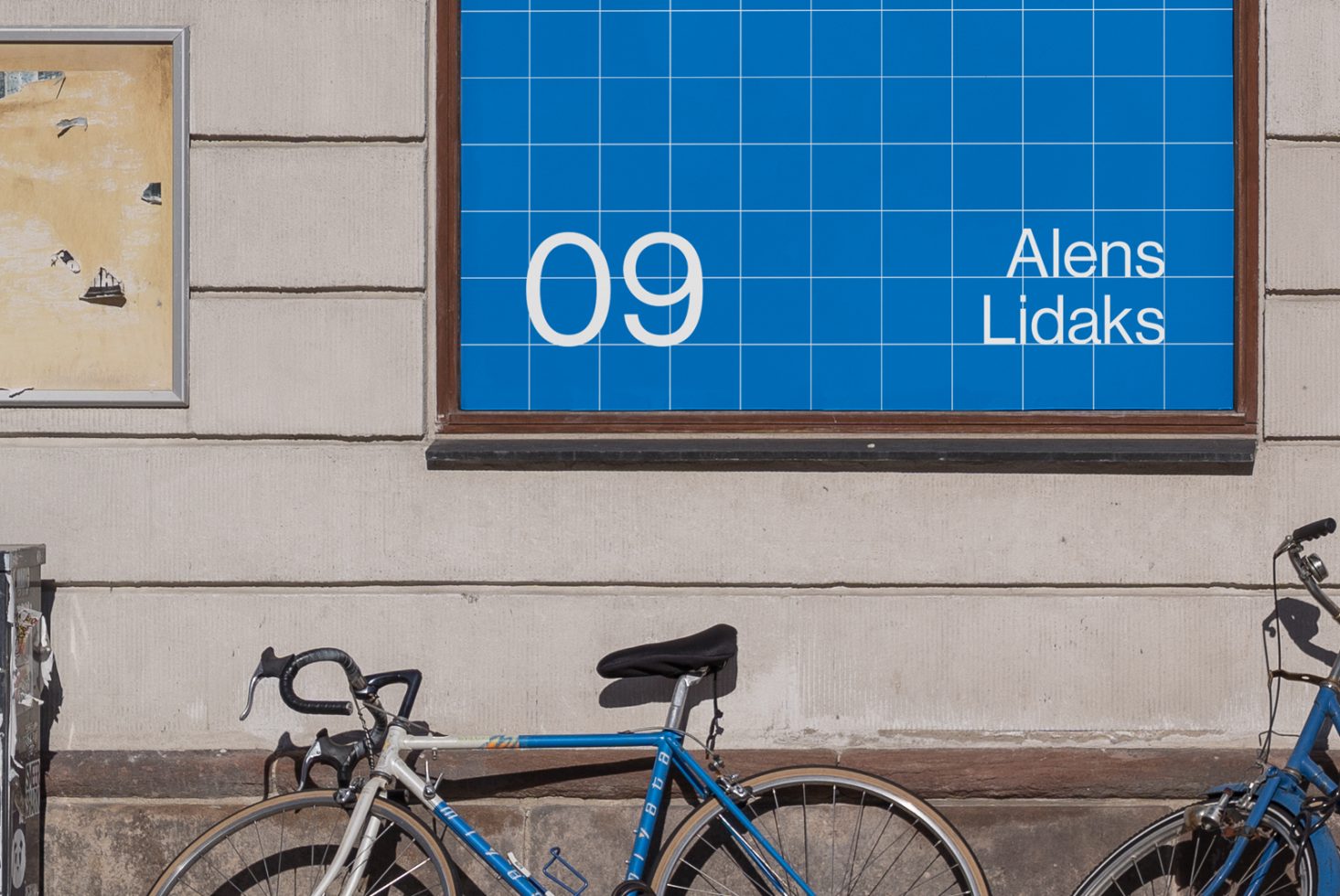 Outdoor wall with grid-based poster design mockup showcasing a blue background and white text bicycles parked nearby urban setting design assets for creators