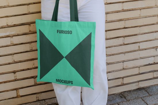 Green tote bag mockup featuring a geometric design worn by a person in white pants against a beige brick wall. Perfect for showcasing branding.