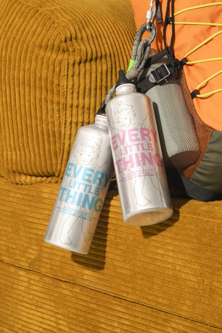 Stainless steel water bottles with custom typography hanging from a backpack on a mustard corduroy background. Perfect for showcasing branding mockups.