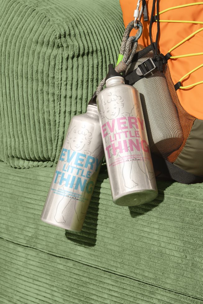 Two metal water bottles with Every Little Thing text and cartoon design strapped to an orange backpack on a green fabric chair. Suitable for mockups or graphics.