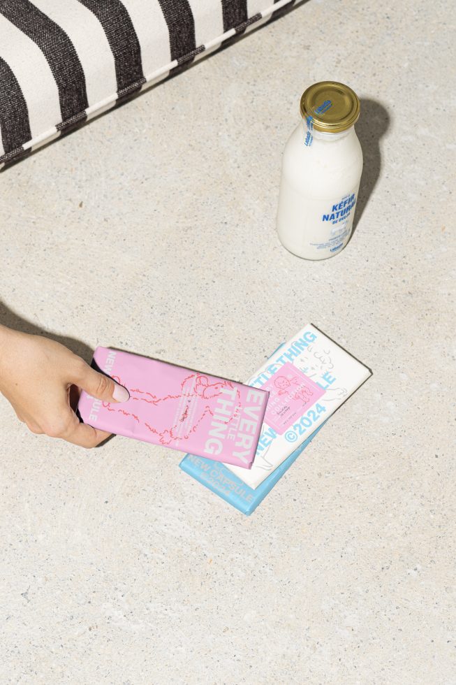 Hand holding pink graphic design booklet on textured surface with striped fabric and bottle of kefir milk in the background mockups templates design resources.