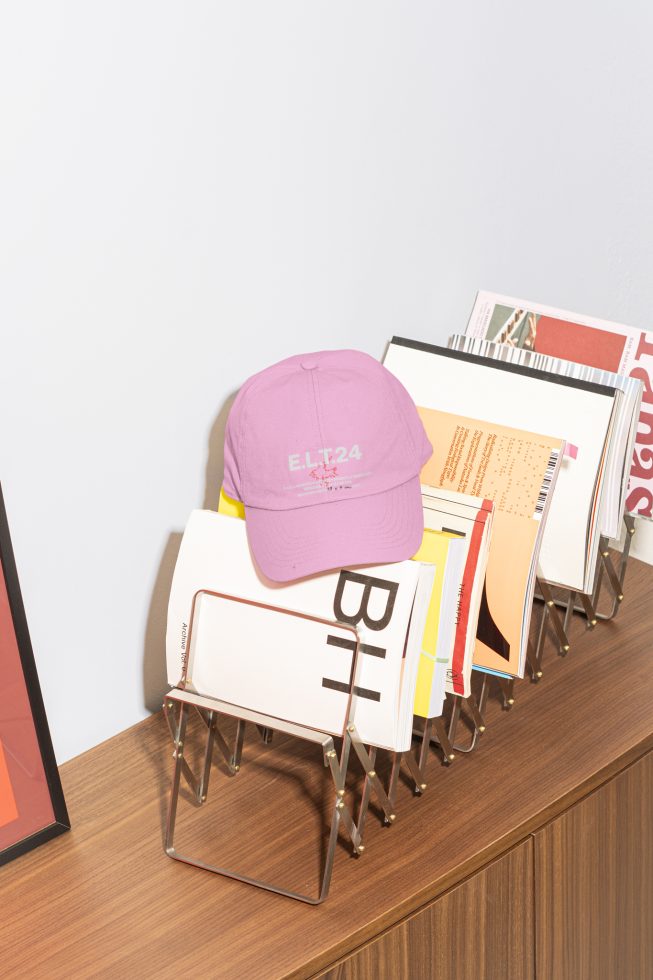 A pink cap placed on a magazine rack filled with various colorful design publications on a wooden table. Suitable for graphics or mockups in designers' marketplace.