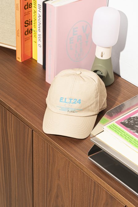 Beige E.L.T.24 baseball cap placed on a wooden shelf with colorful books and a small modern lamp. Ideal for product mockups and graphic design projects.