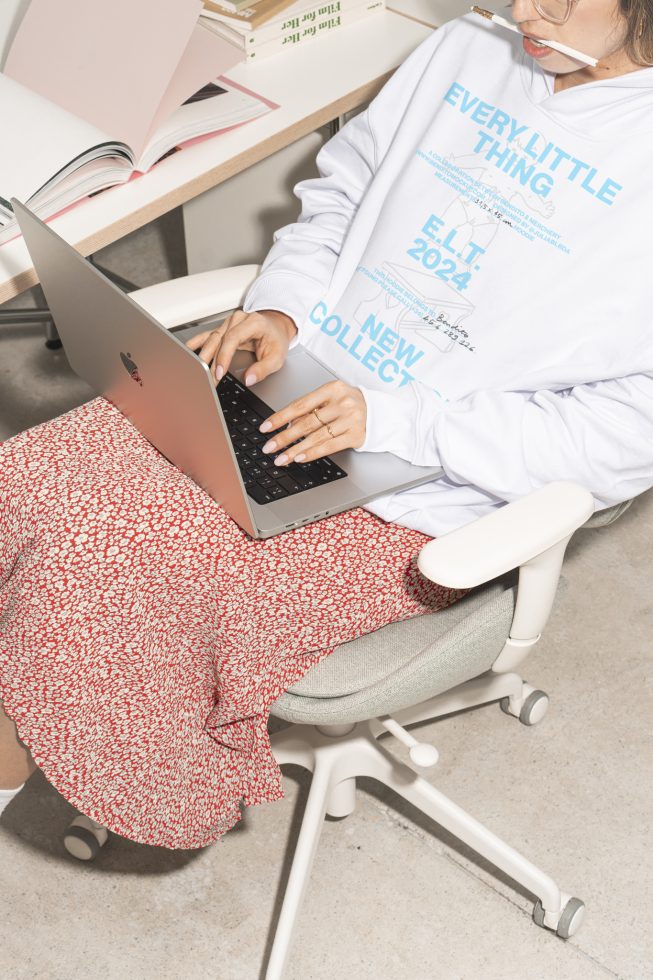 Designer working on laptop in ergonomic chair wearing graphic hoodie and red floral skirt beside desk with open books. Mockup Template Fashion Design Graphic