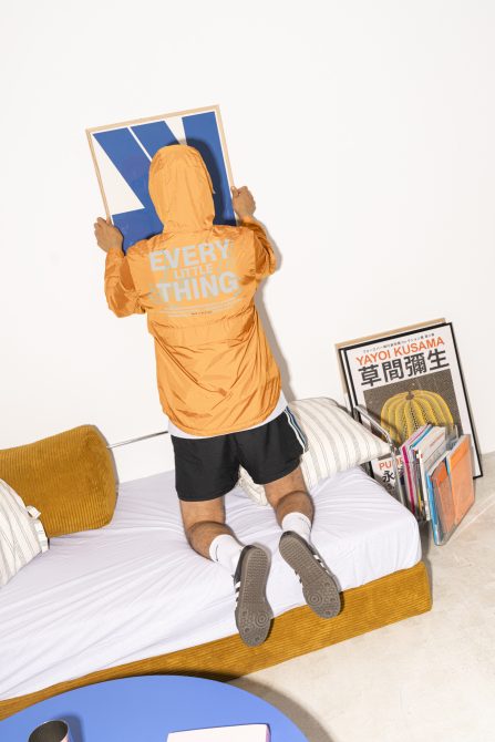 Man in orange jacket hanging blue minimalist print on wall standing on bed with yellow pillow in modern room with books and magazines near bed.