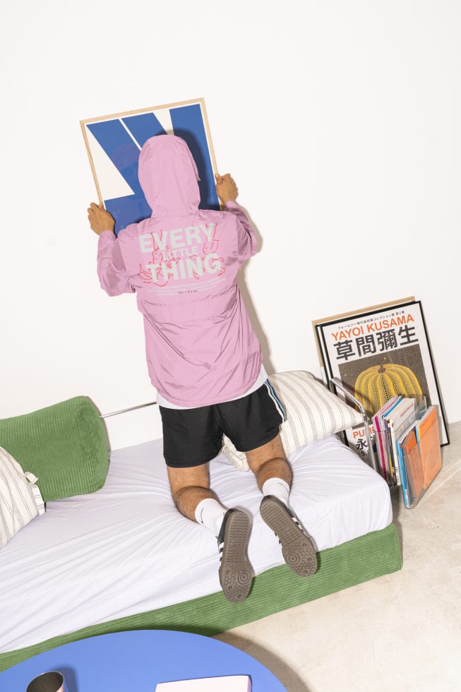 Designer hanging minimalist blue white framed artwork on wall behind green cushion couch while wearing pink jacket black shorts. Books and posters adjacent.