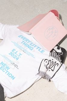 Overhead flat lay of white tee with blue event text, pink box, patterned socks on concrete ground
