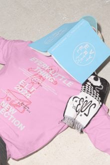 Pink tee with playful line art, blue bag with circular logo, black white socks on concrete ground