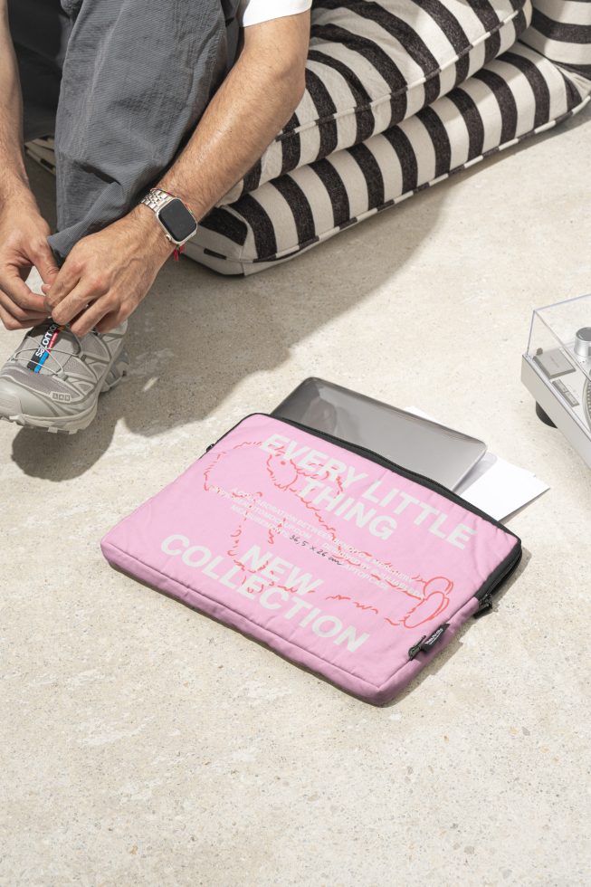 Designer sitting on cushions while tying shoelaces pink laptop sleeve on floor with text every little thing new collection mockup template digital asset