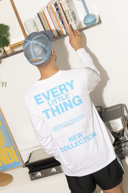 Guy in white long-sleeve tee with blue text reaching to shelf, blue mesh cap