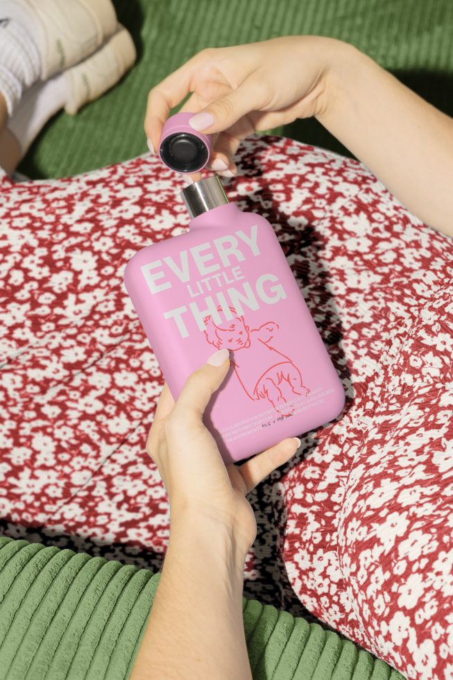 Mockup of a person holding a pink bottle labeled Every Little Thing while sitting on a floral dress, suitable for graphic designers seeking design assets.