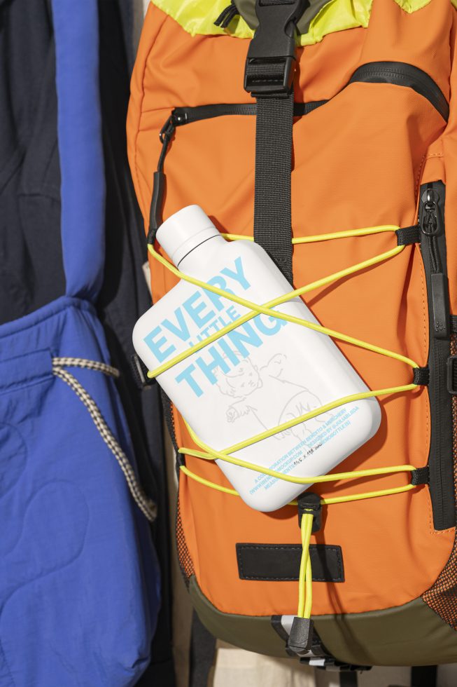 Orange backpack with yellow bungee cord holding white bottle printed with Every Little Thing text hanging next to a blue garment. Ideal for mockups templates.