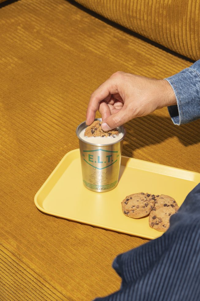 Hand dipping cookie into milk in stainless cup on yellow tray with cookies. Ideal for mockup, graphics, templates. Cozy and inviting design.