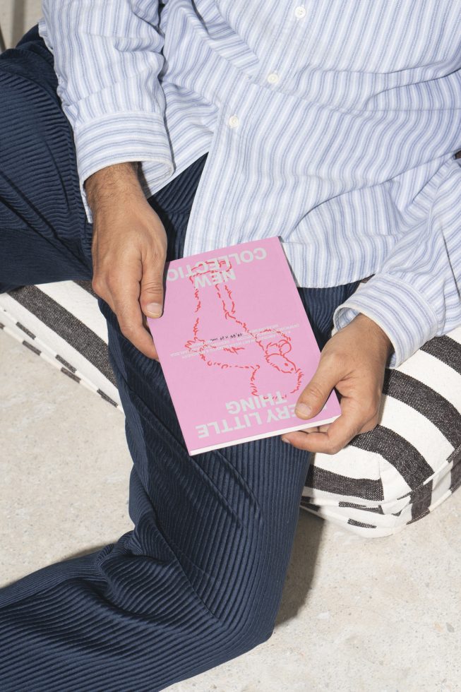 Man holding a pink booklet with NEW COLLECTION text dressed in striped shirt and blue corduroy pants perfect for magazine mockups and print design assets