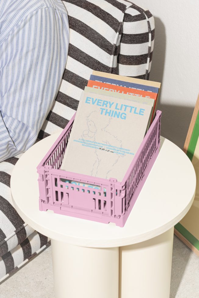 Pink plastic crate with magazines placed on a round white table against striped cushion background. Suitable for mockups and graphic design assets.