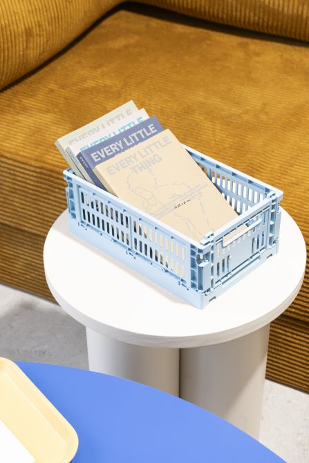 Design mockup of pastel books in a light blue crate on a minimalist white table next to a mustard yellow couch and blue coffee table.