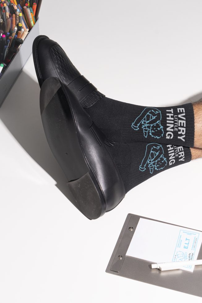 Close-up of a foot in black dress shoe wearing black socks with vintage design, next to metal pencil holder full of pencils and a clipboard with paper. Fashion Mockup.