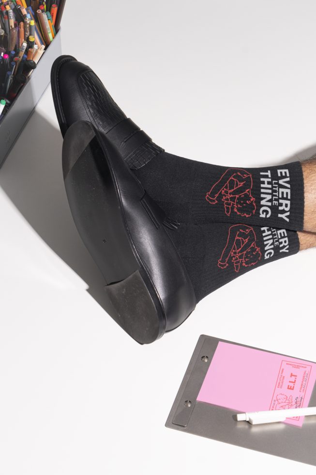 Black leather shoe with black sock featuring red graphic design next to a pencil holder and clipboard with pink paper. Ideal for mockup designs.