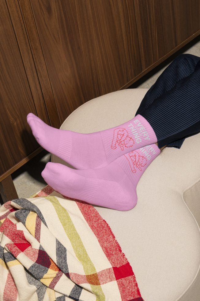 Person wearing pink socks with illustrated design relaxing on beige chair with colorful blanket. Keywords: digital assets, mockup, socks, textile design, designers.