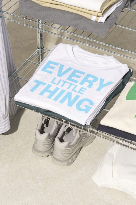 Folded t-shirt reading every little thing on a wire shelf with other clothing and sneakers. Use this t-shirt mockup for apparel design presentations: Keywords: mockup t-shirt apparel design presentation.