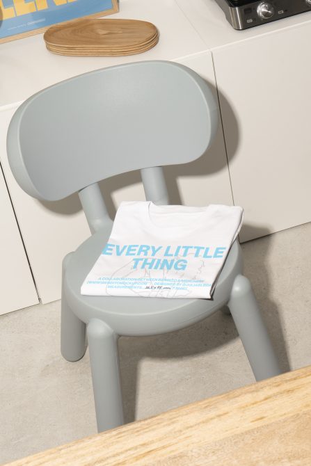 Mockup of a folded white t-shirt with blue text reading Every Little Thing placed on a light gray chair near a table in a minimalist room. Suitable for designers.